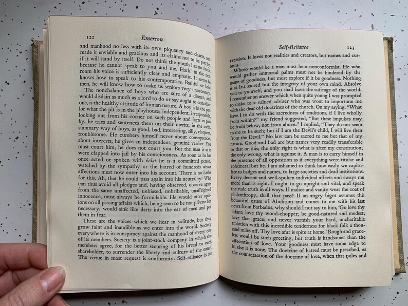 May include: Open book with text on the pages. The left page has the title "Emerson" at the top, and the right page has the title "Self-Reliance". The book is open to pages 122 and 123.