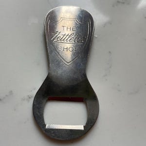 May include: A vintage silver bottle opener with the text "THE Nettleton SHOE" in a shield design. The opener has a curved handle and a rectangular opening for removing bottle caps. The metal has a worn, aged appearance.