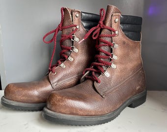 May include: A pair of brown leather work boots with red laces. The boots have a rugged, worn appearance, with a black padded collar and a thick, dark sole. The laces are threaded through metal eyelets.