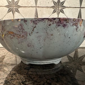 May include: A large, decorative bowl with a light blue and purple marbled design. The rim has a gold trim. The bowl sits on a small, white base. The bowl is displayed on a dark, speckled surface.