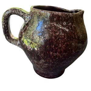 Può includere: Una brocca in ceramica con corpo arrotondato e un manico singolo. La superficie presenta una smaltatura screziata con toni marrone scuro, viola scuro e macchie verde muschio, accentuata da una texture bianca punteggiata su tutto l'esterno.