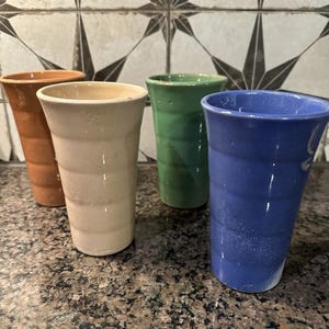 Peut inclure: Quatre gobelets en céramique faits à la main, dans les tons de marron, crème, vert et bleu. Les gobelets ont une forme légèrement conique et une surface texturée. L'arrière-plan présente un motif de carreaux décoratifs. Parfaits pour servir des boissons.