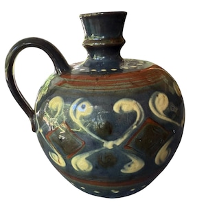 May include: A round ceramic pitcher with a dark blue glaze, featuring a single handle and a narrow neck. The body is decorated with cream-coloured swirling patterns and brown diamond shapes, with small white dots encircling the base and neck.