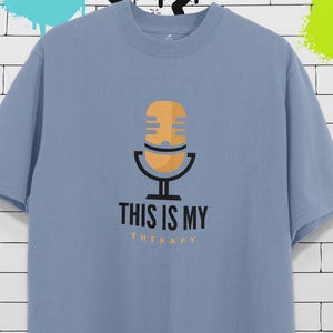 Puede incluir: Camiseta azul claro con un gráfico de un micrófono naranja y negro. El texto "THIS IS MY THERAPY" está impreso debajo en negro y naranja. La camiseta tiene mangas cortas y cuello redondo.