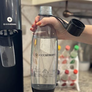Może przedstawiać: Przezroczysta butelka SodaStream z czarną podstawą i zakrętką trzymana jest w pobliżu czarnej maszyny SodaStream. Butelka jest wypełniona przezroczystym płynem. Widoczne jest logo SodaStream.