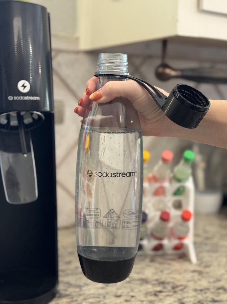 Może przedstawiać: Przezroczysta butelka SodaStream z czarną podstawą i zakrętką trzymana jest w pobliżu czarnej maszyny SodaStream. Butelka jest wypełniona przezroczystym płynem. Widoczne jest logo SodaStream.