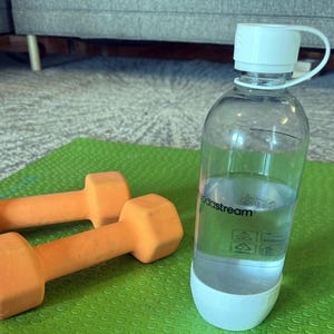 Może przedstawiać: Przezroczysta butelka na wodę SodaStream z białą zakrętką i podstawą, wypełniona wodą, stoi na zielonej macie do ćwiczeń. Dwie pomarańczowe hantle leżą obok butelki. Scena sugeruje skupienie na fitnessie i nawodnieniu.