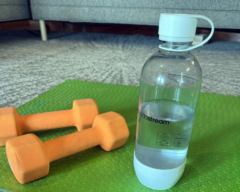 Może przedstawiać: Przezroczysta butelka na wodę SodaStream z białą zakrętką i podstawą, wypełniona wodą, stoi na zielonej macie do ćwiczeń. Dwie pomarańczowe hantle leżą obok butelki. Scena sugeruje skupienie na fitnessie i nawodnieniu.