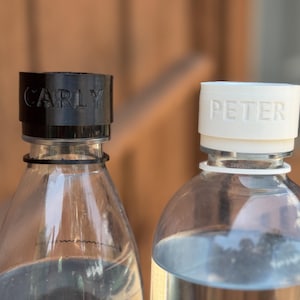 Może przedstawiać: Dwie przezroczyste butelki z wodą, każda z personalizowaną nakrętką. Jedna nakrętka jest czarna i ma napis "CARLY", druga jest biała i ma napis "PETER". Butelki na rozmytym tle drewnianym.