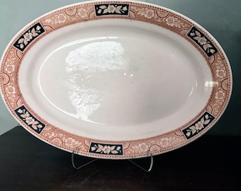 May include: An oval ceramic serving platter with a white center and a decorative border. The rim features a repeating pattern of light brown floral motifs and geometric shapes interspersed with dark blue rectangular floral panels.