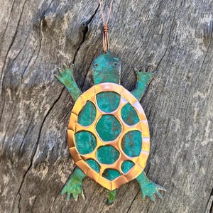 May include: A copper turtle ornament with a green patina finish. The turtle's shell is cut out and has a copper finish. The ornament has a loop at the top for hanging.