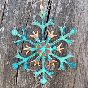 Puede incluir: Un adorno con forma de copo de nieve que presenta un diseño en capas con un acabado de pátina verde azulado oxidado y desgastado en la estructura exterior y detalles de metal pulido color cobre en el núcleo estrellado y el disco central.