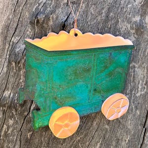 May include: A decorative ornament shaped like a vintage train car. The train car is a weathered green with orange wheels and a copper-colored roof. A copper wire is attached for hanging. The ornament is set against a rustic wooden background.