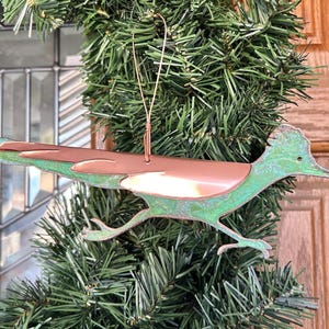 Può includere: Un ornamento di roadrunner verde e rame con un gancio di filo. L'ornamento è in metallo e ha una finitura invecchiata.
