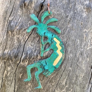 May include: A turquoise green Kokopelli figurine ornament with a yellow zigzag pattern. The ornament is made of metal and has a rustic finish.