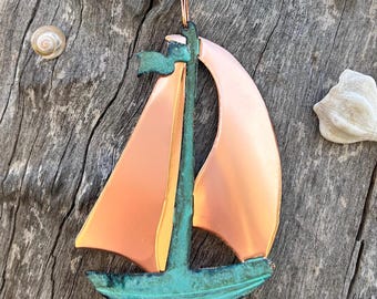 Pode incluir: Um ornamento de veleiro de cobre com uma vela verde e um cabide de arame de cobre. O ornamento está pendurado em uma superfície de madeira.