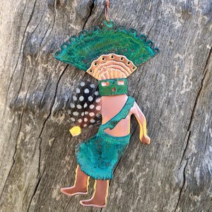 May include: A copper and green metal ornament depicting a stylized figure with a feathered headdress and a fan. The figure is wearing a green skirt and has a copper face mask.