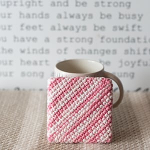 May include: A square, hand-crocheted coaster with a diagonal pattern of alternating pink and white stripes. A beige mug sits behind the coaster, and the background features inspirational text.