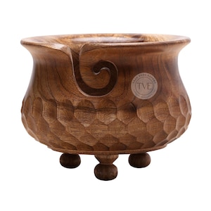 May include: A wooden yarn bowl with a carved spiral yarn guide. The bowl features a textured, honeycomb pattern and stands on three small, rounded feet. A circular logo with the text "TRAD VEDA EXPORT" is visible. The bowl is a warm brown colour.