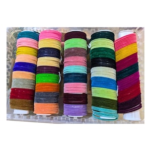 Puede incluir: Una colección de pulseras coloridas en un recipiente de plástico transparente. Las pulseras están apiladas en columnas, mostrando una variedad de colores, incluyendo rosa, amarillo, verde, azul y morado. La imagen resalta los tonos vibrantes y las texturas de las joyas.