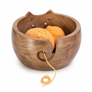 Puede incluir: Un cuenco de madera de color marrón oscuro con un borde en forma de gato, dos orejas puntiagudas y pequeños recortes circulares. Una abertura en espiral en la parte frontal permite que un hilo de lana naranja pase desde dos ovillos naranjas.