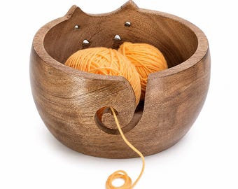 May include: A round, dark brown wooden yarn bowl featuring a cat-shaped rim with two pointed ears and small circular cutouts. A spiral-shaped opening on the front allows a single strand of orange yarn to feed through from two orange yarn balls inside.