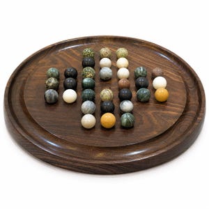 Peut inclure: Un plateau circulaire en bois brun foncé avec un rebord surélevé, présentant une collection de sphères en pierre polie disposées en forme de croix. Les sphères présentent des couleurs variées, notamment blanc, noir, vert, gris et beige.