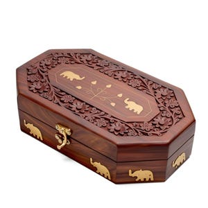 Puede incluir: Una caja de madera marrón oscuro con forma octogonal. La tapa presenta patrones florales tallados y detalles dorados de elefantes y flores. Un cierre dorado asegura la caja. La caja probablemente se utiliza para guardar joyas o objetos pequeños.