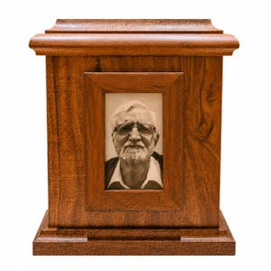 May include: A rectangular wooden cremation urn with a polished medium-brown finish and a prominent grain pattern. The front features a centered, vertical rectangular frame containing a black and white portrait of an older person with a beard and glasses.