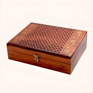 Puede incluir: Una caja rectangular de madera con tapa abatible que presenta un patrón de celosía cortado con láser en el centro e incrustaciones florales de latón en los bordes. La caja tiene un acabado pulido marrón rojizo y un cierre de latón.
