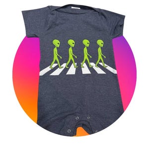 May include: Dark gray baby onesie featuring a graphic of four green aliens walking across a white crosswalk. The onesie has short sleeves and snap closures at the bottom. The design is set against a colorful, circular background.