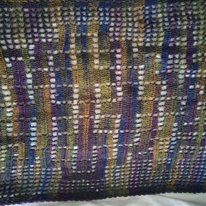 May include: A handmade, open-weave textile in a variety of colors. The piece features a repeating pattern of vertical stripes in shades of blue, purple, green, and gold. The open weave creates a delicate, airy texture, suitable for a shawl or scarf.