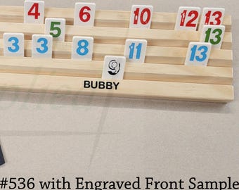 Pode incluir: Um suporte de madeira clara para peças com quatro ranhuras horizontais, contendo várias peças de jogo numeradas em vermelho, verde e azul. A palavra BUBBY está gravada na frente. Ao lado, há uma bolsa azul-escura com cordão e o texto Simplified Woodcraft Products, LLC.