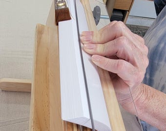 Pode incluir: Uma prensa de encadernação de madeira com uma pilha de papel branco sendo pressionada. A prensa tem um guia de acrílico transparente e uma alça de madeira. A prensa está sobre uma base de madeira com uma superfície de tecido.
