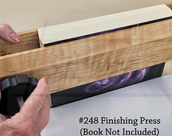 Pode incluir: Uma prensa de acabamento de madeira com botões pretos e uma haste prateada. A prensa está segurando um livro com uma capa roxa e preta. O texto na imagem diz "#248 Finishing Press (Book Not Included)".