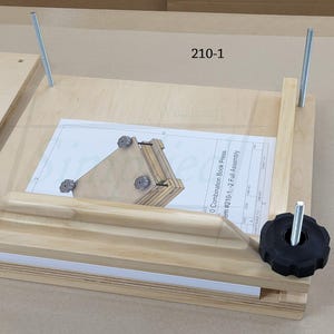 以下が含まれることがあります： 2本の垂直な金属製ネジ棒と2つの黒い星型プラスチックノブを備えた明るい色の木製ブックプレス。ベースには「210-1」とラベル付けされた白い説明書が置かれており、組み立て図が示されています。