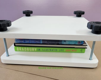 Pode incluir: Prensa de livros branca com quatro botões pretos e duas hastes rosqueadas prateadas. A prensa está segurando uma pilha de três livros, incluindo "Bradbury: An Illustrated Life" de Jerry Weist. "Livros não incluídos" está escrito em verde abaixo da prensa.