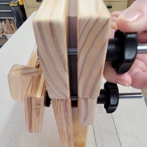 May include: A wooden workbench with two clamps and a metal screw. The workbench is made of light wood and has a natural finish. The clamps are black and have a knob on the top. The screw is silver and has a threaded shaft.