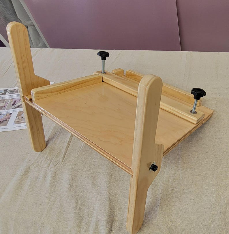 May include: A wooden easel with two adjustable legs and two black knobs. The easel is made of light wood and has a flat surface.