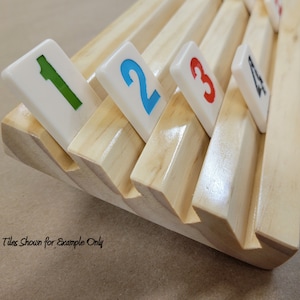 May include: Wooden rack with slots for holding numbered tiles. The rack is made of light wood and has a natural finish. The tiles are white with colored numbers. The tiles shown are numbered 1, 2, 3, and 4. "Tiles Shown for Example Only" is written on the bottom of the rack.