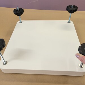 May include: White square platform with four black knobs on adjustable metal legs. The knobs are used to level the platform.