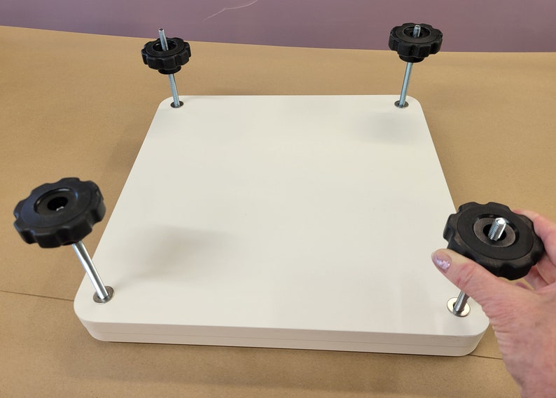 May include: White square platform with four black knobs on adjustable metal legs. The knobs are used to level the platform.