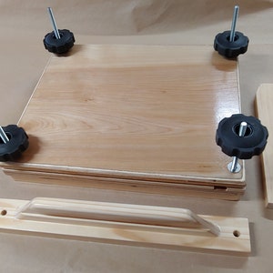 May include: A light-colored wooden book press featuring a square base and top plate, four vertical metal threaded rods with black plastic star-shaped knobs at each corner, and two separate wooden handle components positioned nearby.
