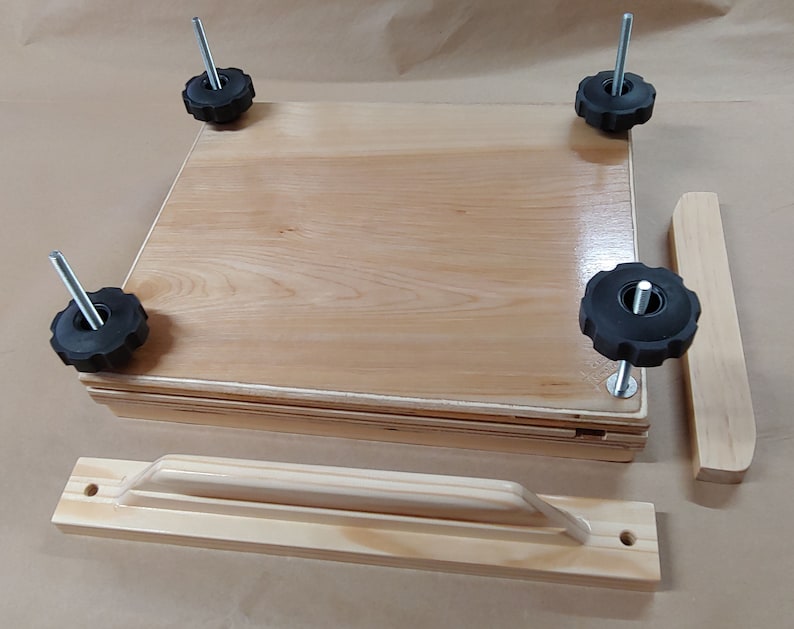 May include: A light-colored wooden book press featuring a square base and top plate, four vertical metal threaded rods with black plastic star-shaped knobs at each corner, and two separate wooden handle components positioned nearby.