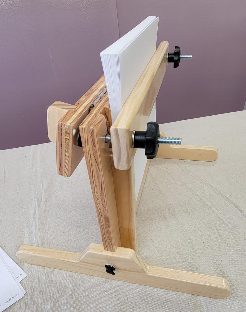May include: A wooden book press with two adjustable clamps and a base with three legs. The press is designed to hold a stack of paper in place while it is being pressed.