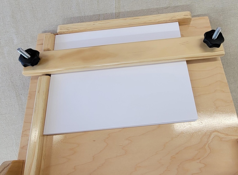 May include: A wooden printing press with a white sheet of paper clamped in place. The press has two wooden knobs on either side that are used to tighten the clamps.
