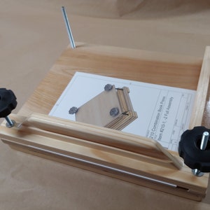 May include: A light-colored wooden book press featuring two vertical metal threaded rods with black plastic star-shaped knobs. A white instruction sheet titled 210 Combination Book Press Item #210-1, -2 Full Assembly rests on the wooden base.