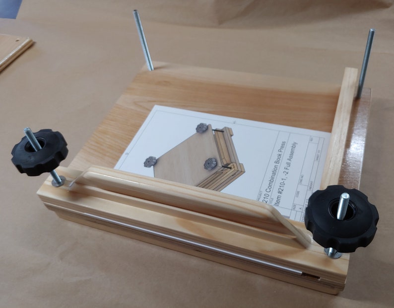 May include: A light-colored wooden book press featuring two vertical metal threaded rods with black plastic star-shaped knobs. A white instruction sheet titled 210 Combination Book Press Item #210-1, -2 Full Assembly rests on the wooden base.
