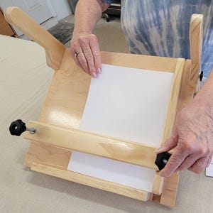 May include: A wooden bookbinding press with a white piece of paper clamped in place. The press has two wooden arms that can be tightened with black knobs.