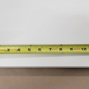 May include: A white shelf with a yellow tape measure showing a measurement of 11 1/2 inches. The shelf has two silver metal brackets attached to the underside.